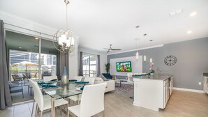 Cozinha moderna com ilha central, área de jantar com mesa de vidro e sala de estar com TV e assentos.