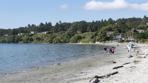 A beach with people walking along the shore, a boat on the water, and houses in the background. - 10 Mile Point Surfside Suite (Victoria)