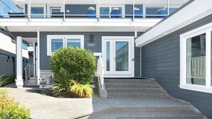 A two-story building with a grey exterior, a white door, and a small garden with a bush and fern. - 10 Mile Point Surfside Suite (Victoria)