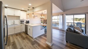 A modern kitchen with white cabinets, a central island, and pendant lighting. There is a dining area with a table and chairs, and a balcony with a view.