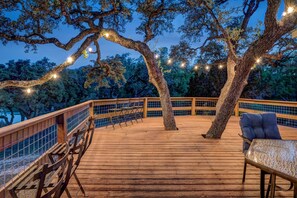 Una terraza de madera con baranda, una mesa y sillas, rodeada de árboles y luces colgantes.