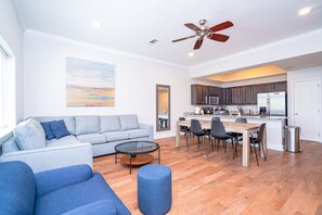 A modern living room with a sofa, coffee table, and ottoman, a dining area with chairs and a table, a kitchen with appliances, and a ceiling fan.