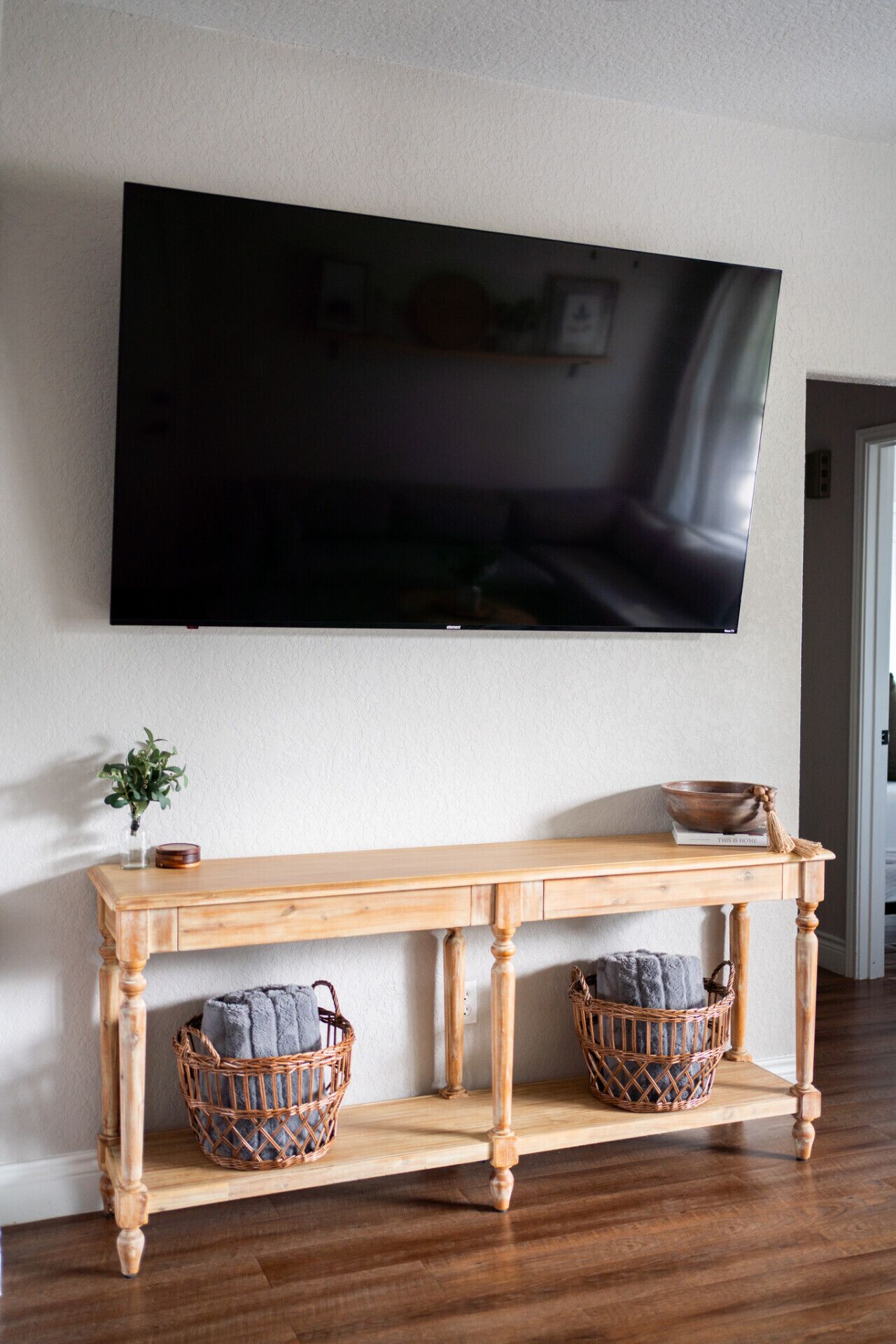 Une console en bois, agrémentée de paniers en osier et d’un saladier, est placée devant un téléviseur à écran plat fixé au mur.