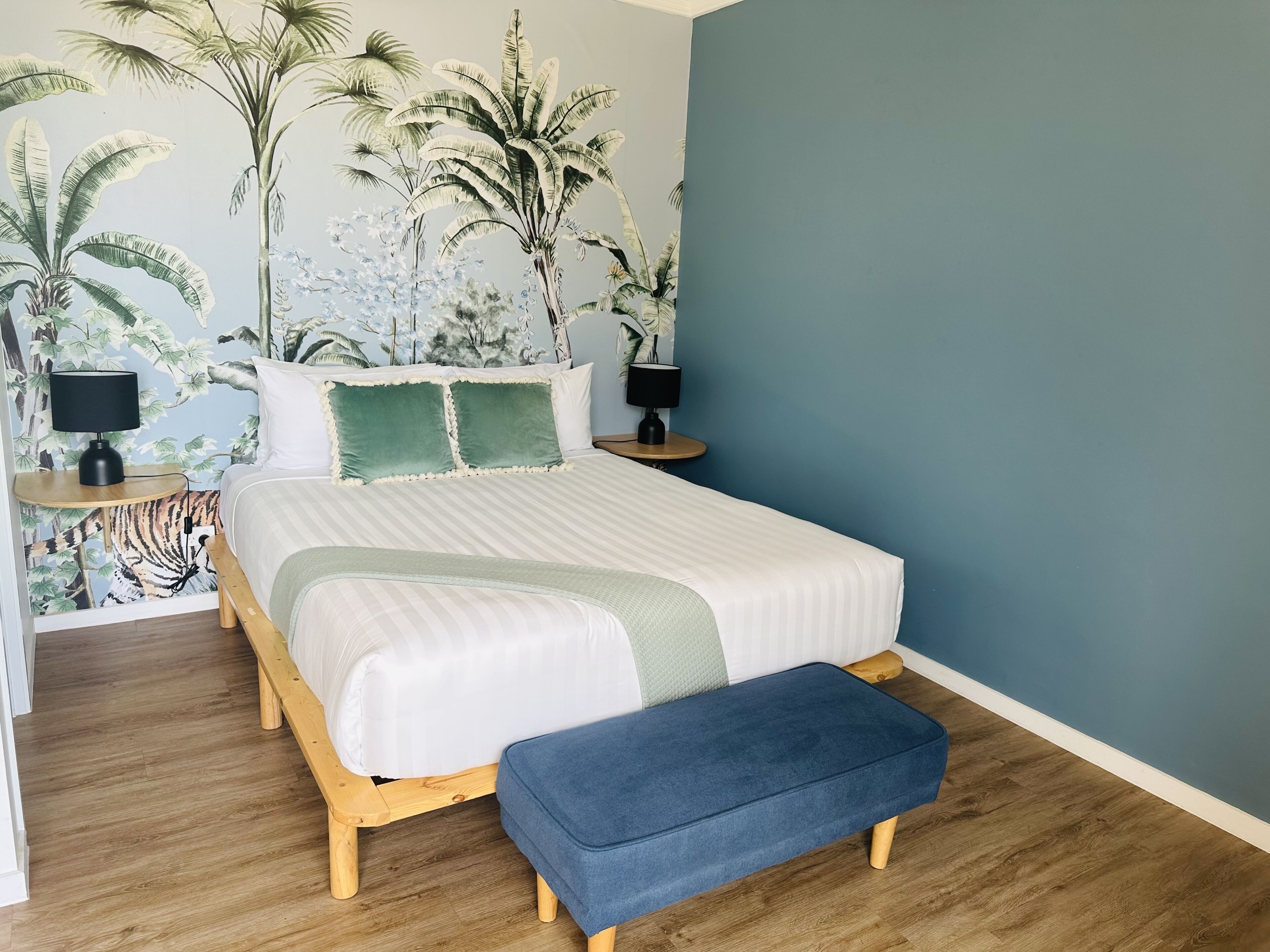 A bedroom with a bed, two bedside tables, a bench, and a wall with tropical wallpaper.