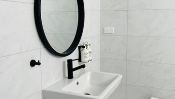 Un baño con un lavabo blanco, grifo negro y un espejo redondo con marco negro. Hay un taburete de madera con una toalla blanca encima.