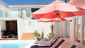 A poolside area with lounge chairs and a table, a red umbrella, and a glass building in the background.