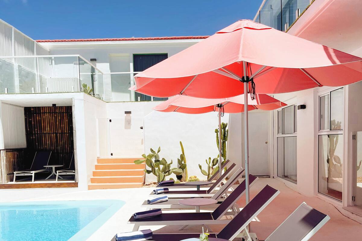 A poolside area with lounge chairs and a table, a red umbrella, and a glass building in the background.