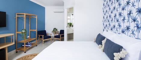 A bedroom with a bed, a wooden shelf unit, two chairs, a small table, and a wall with a palm tree pattern.