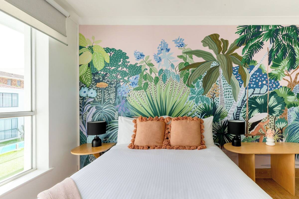 A bedroom with a large bed, two nightstands, a window, and a tropical-themed mural on the wall.