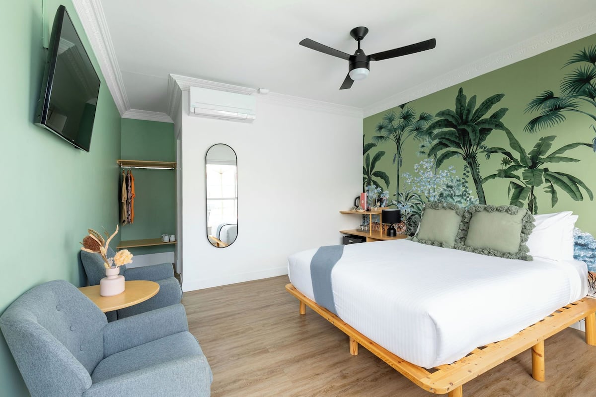 A bedroom with a bed, a chair, a TV, a ceiling fan, and a wall with a tropical mural.