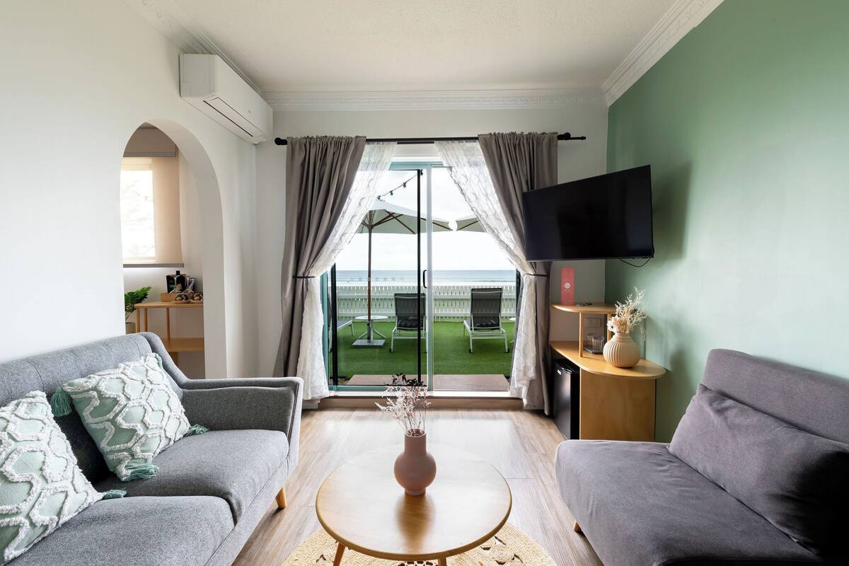 A living room with a sofa, a coffee table, and a TV. There is a window with curtains and a view of a deck.