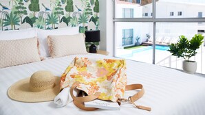 A hotel room with a bed, a floral-patterned bag, a straw hat, and a view of a pool and buildings outside.