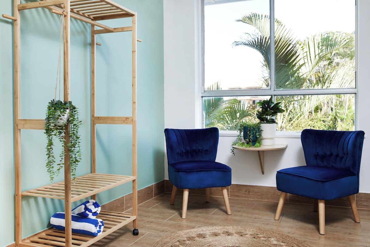 A room with a wooden shelving unit, two blue velvet chairs, and a window with a view of palm trees.
