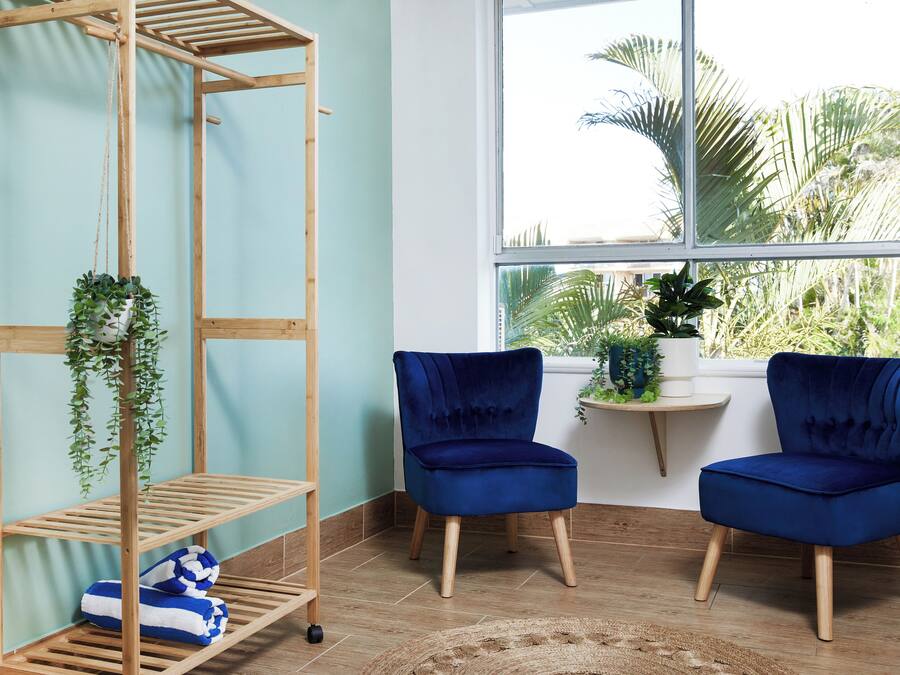 A room with a wooden shelving unit, two blue velvet chairs, and a window with a view of palm trees.