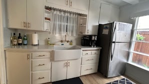 Cocina compacta con armarios blancos, refrigerador de acero inoxidable, cafetera y ventana con vista a una cerca de madera.