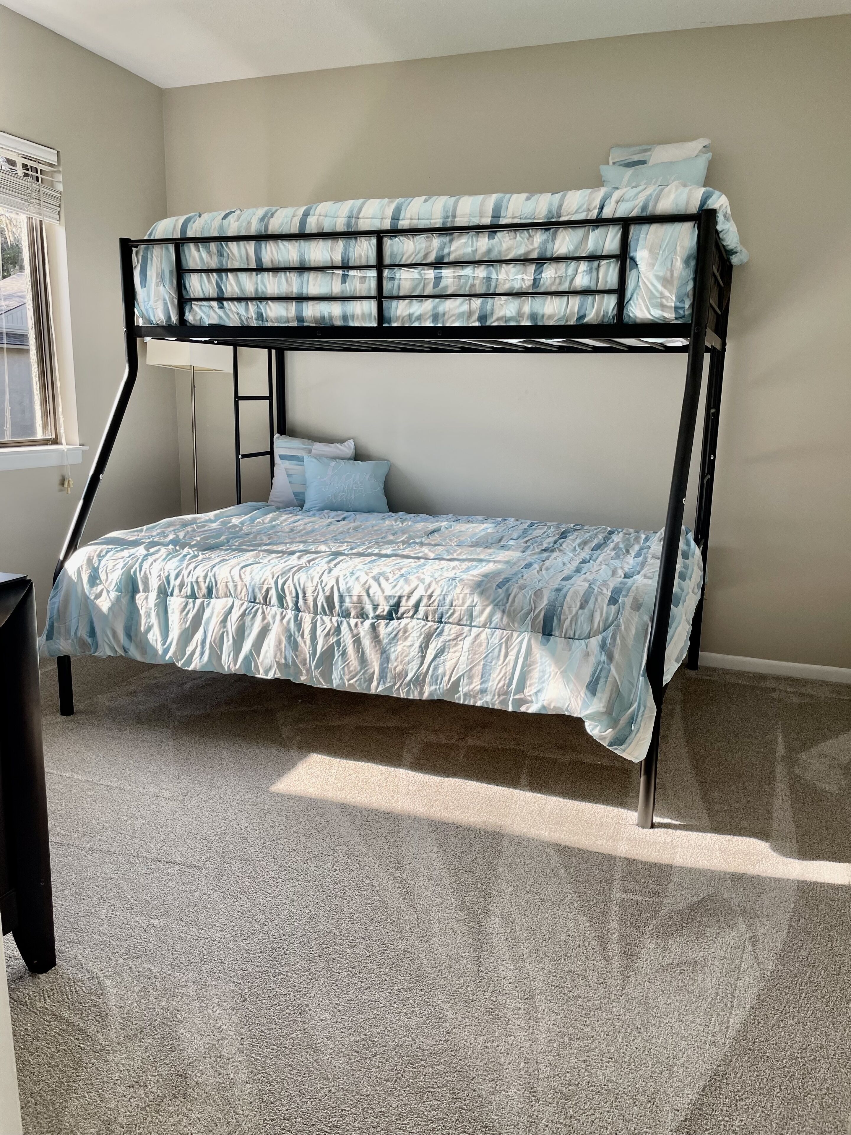 Un lit superposé avec une literie à rayures bleues et blanches, une fenêtre avec des stores et un sol recouvert d’une moquette.