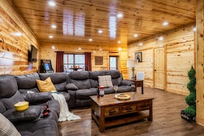 Ein gemütliches Holzhaus-Interieur mit einem ledernen Ecksofa, einem Couchtisch aus Holz und einem an der Wand befestigten Fernseher.