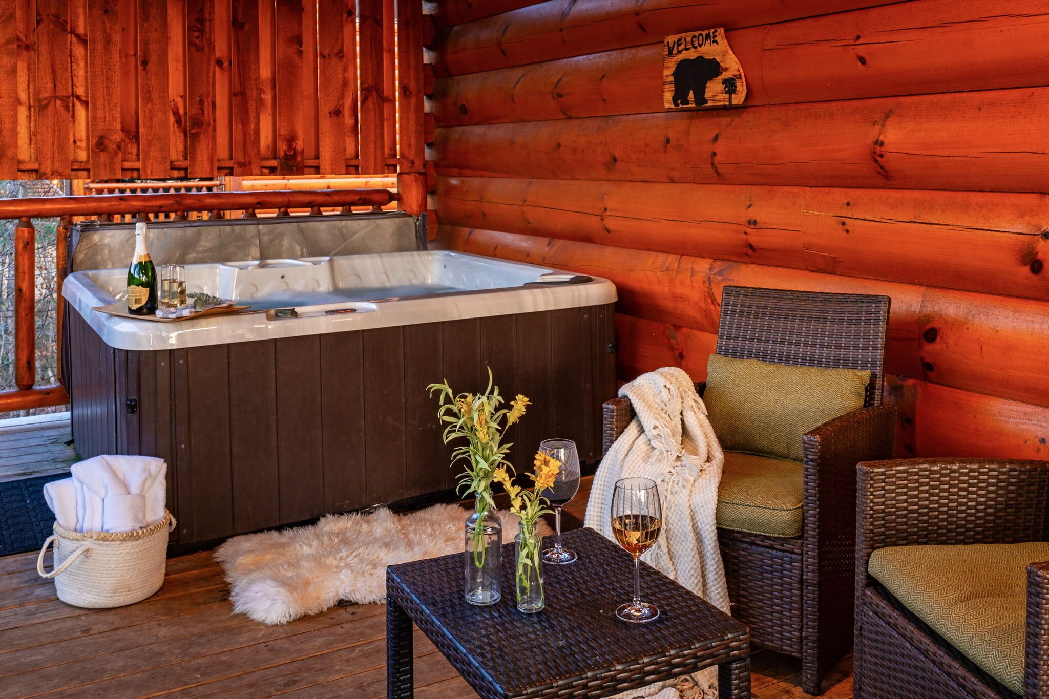 Interior acolhedor de uma cabana com hidromassagem, cadeiras de vime, uma mesa de vidro e copos de vinho, e um letreiro com a palavra 'Bem-vindo'.