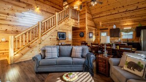Ein gemütliches Blockhaushaus-Interieur mit einer Holztreppe, einem grauen Sofa, einem Essbereich und einer Küche mit Kühlschrank.