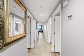 A hotel hallway with white walls, a tiled floor, and a large ornate mirror on the left.