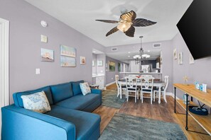 A living room with a blue sofa, a dining area with a white table and chairs, a ceiling fan, and artwork on the walls.