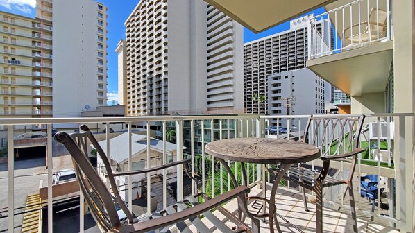 A balcony with a round table and two chairs, overlooking a parking garage and adjacent buildings.