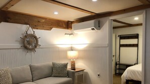 A cozy room with a sofa, a wooden coffee table, a wall-mounted lamp, a ship's wheel decoration, and a bunk bed in the adjacent room. - Top-Rated Vineyard Retreat: Hot Tub + Private Lambert’s Cove Beach Access! (West Tisbury)