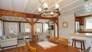A spacious dining area with a wooden table and chairs, a chandelier, and a kitchen in the background. - Top-Rated Vineyard Retreat: Hot Tub + Private Lambert’s Cove Beach Access! (West Tisbury)