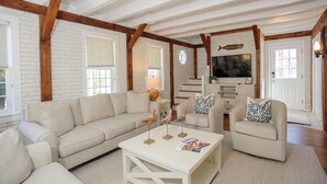 A living room with a beige sofa, a white coffee table, and a television. There are wooden beams on the ceiling and a staircase in the background. - Top-Rated Vineyard Retreat: Hot Tub + Private Lambert’s Cove Beach Access! (West Tisbury)