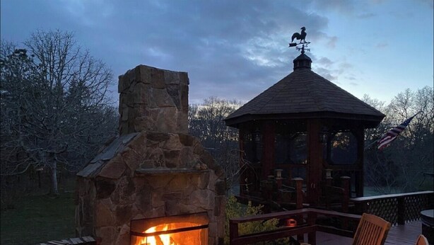 A stone fireplace with a fire, a gazebo, and wooden deck chairs. - Top-Rated Vineyard Retreat: Hot Tub + Private Lambert’s Cove Beach Access! (West Tisbury)