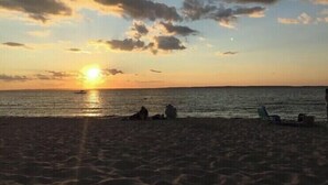 A beach at sunset with the sun low on the horizon, calm sea, and a few people sitting on the sand. - Top-Rated Vineyard Retreat: Hot Tub + Private Lambert’s Cove Beach Access! (West Tisbury)