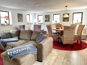 Een moderne woonkamer met een beige bank, rieten stoelen, een eettafel en kunstwerken aan de muur.