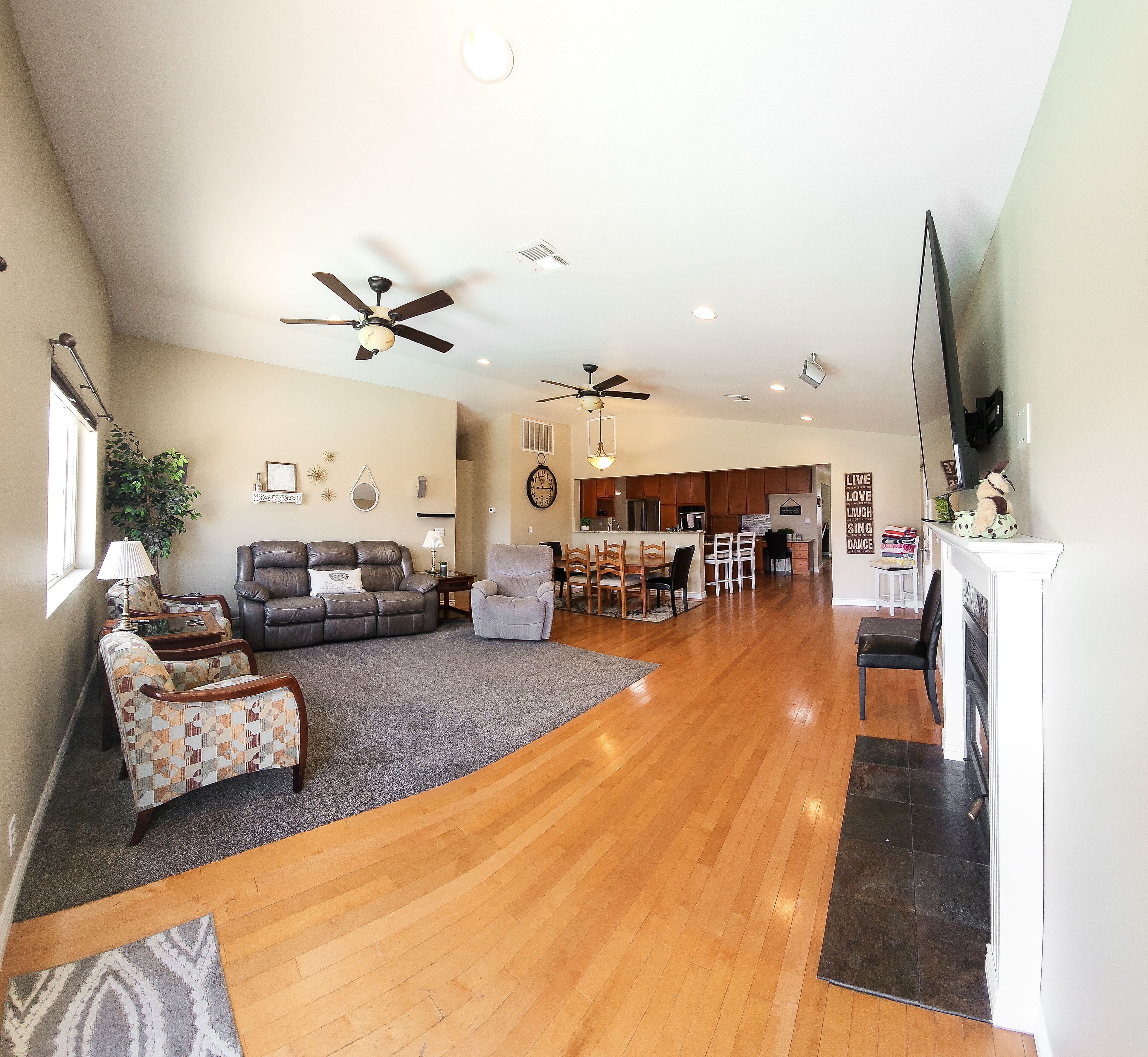 A spacious living area with a wooden floor, a grey carpet, a sofa, a patterned armchair, a dining table with chairs, and a kitchen in the background.