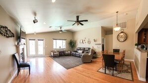 A spacious living area with hardwood floors, a ceiling fan, a dining area with a table and chairs, and a kitchen visible through an open doorway. - Desert Pool Retreat (Mesquite)