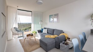 Ein modernes Wohnzimmer mit einem grauen Ecksofa, einem Hängekorbstuhl, einem kleinen runden Tisch und Blick ins Freie durch ein großes Fenster.