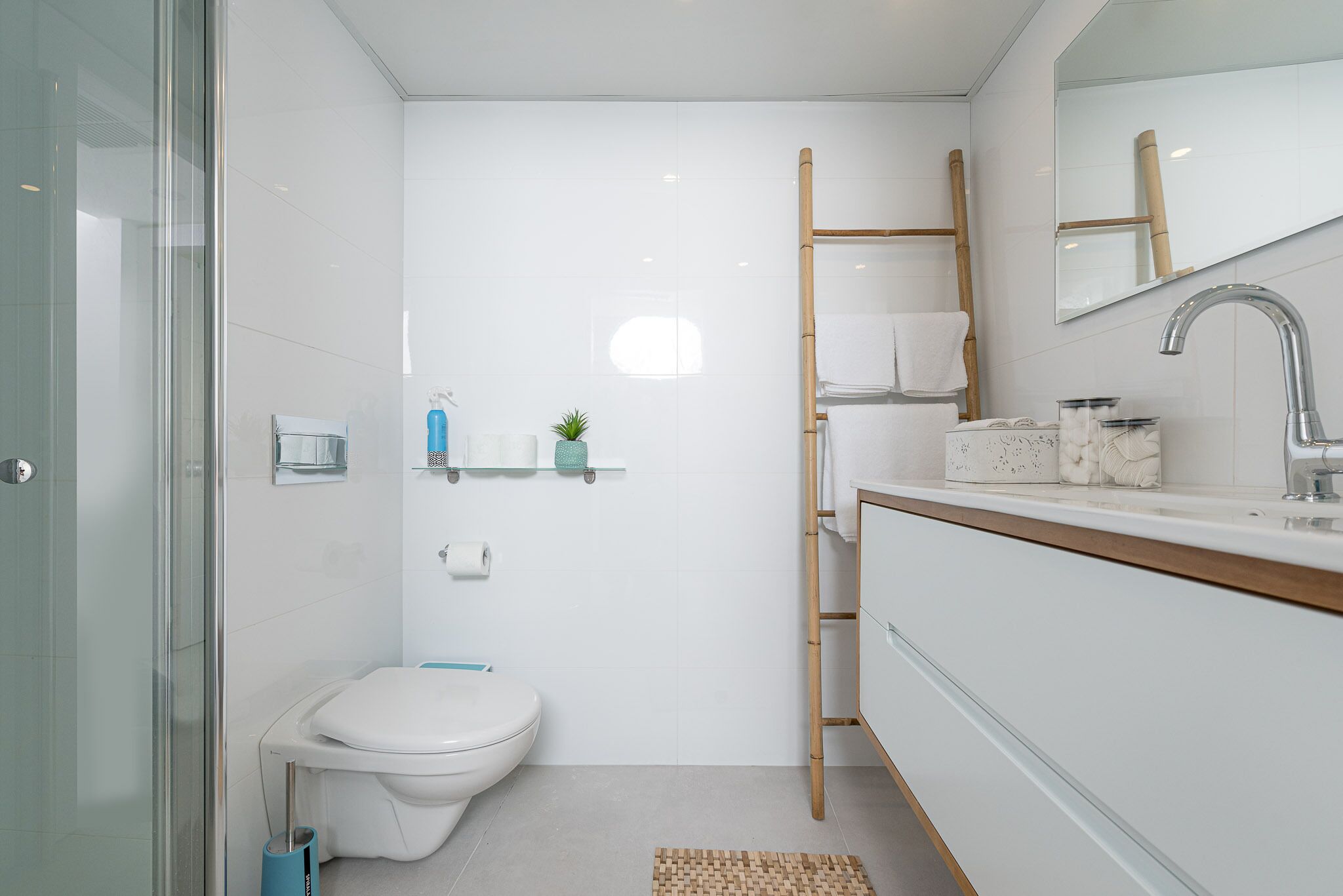 A modern bathroom with a glass shower enclosure, a white toilet, a wooden ladder, a white sink with a mirror, and a small shelf with toiletries.