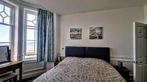 A bedroom with a bed, a desk, a chair, two paintings, and a window with curtains. - With its wonderful, far reaching sea views, this first floor apartment certainly does have the WOW f (Sheringham)