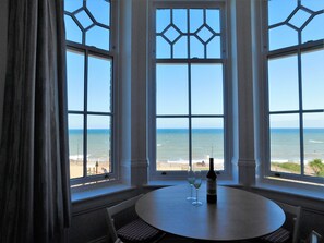 A room with a large window overlooking the ocean, a round table with two wine glasses and a bottle, and a chair with a checkered cushion. - With its wonderful, far reaching sea views, this first floor apartment certainly does have the WOW f (Sheringham)