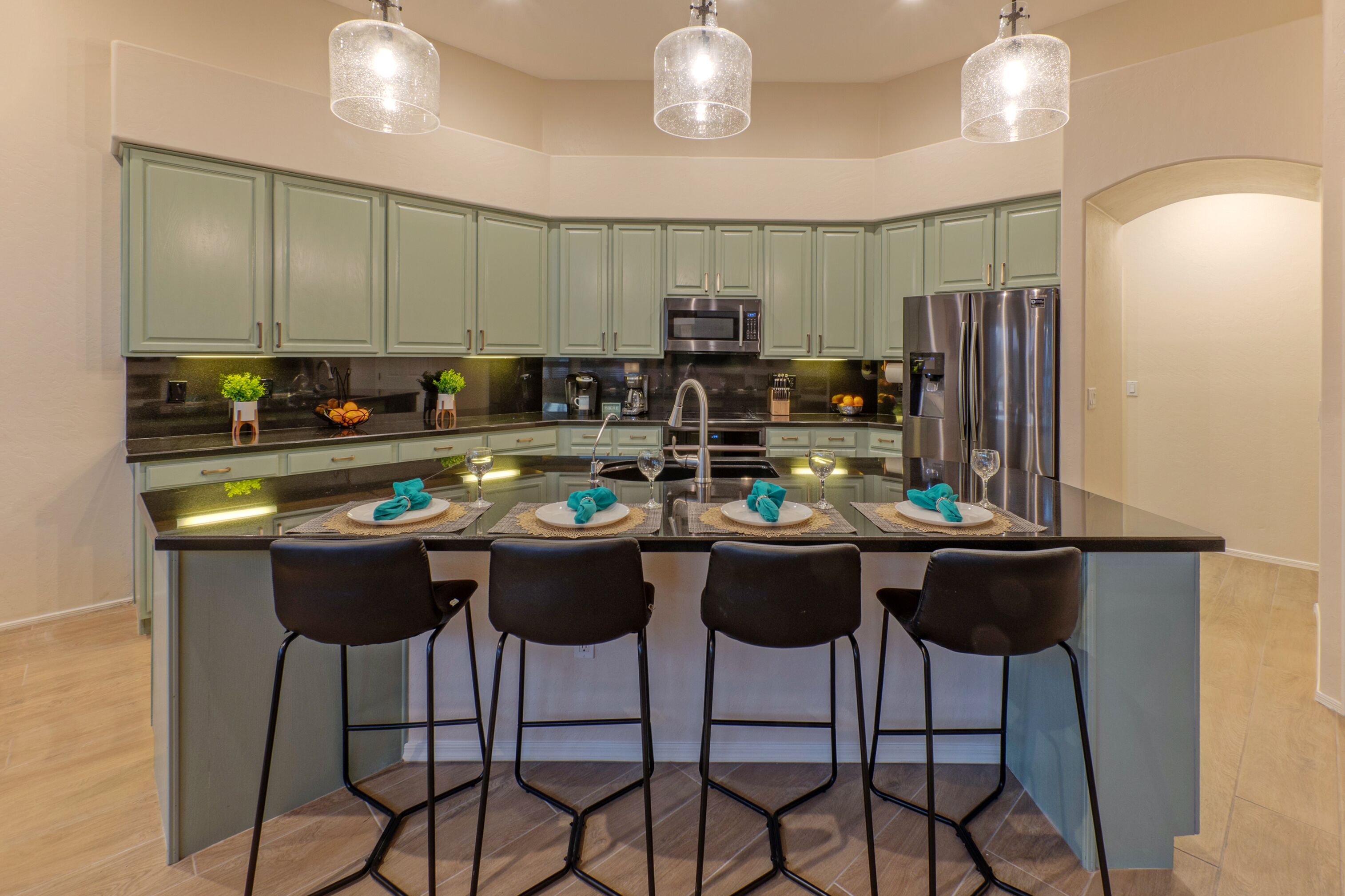 Una cucina moderna con mobili verdi, un'isola centrale con sgabelli da bar, elettrodomestici in acciaio inossidabile e illuminazione a sospensione.