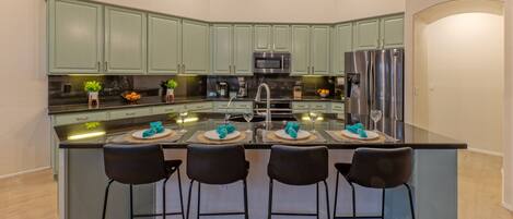 Una cucina moderna con mobili verdi, un'isola centrale con sgabelli da bar, elettrodomestici in acciaio inossidabile e illuminazione a sospensione.
