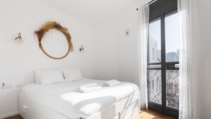 Un lit blanc avec une literie et des serviettes blanches, un plancher en bois, un balcon donnant sur des immeubles et un luminaire fixé au mur.