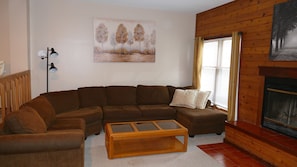 Ein Wohnzimmer mit einem braunen Ecksofa, einem Holztisch, einem Baumgemälde, einer Stehlampe und einem Kamin.