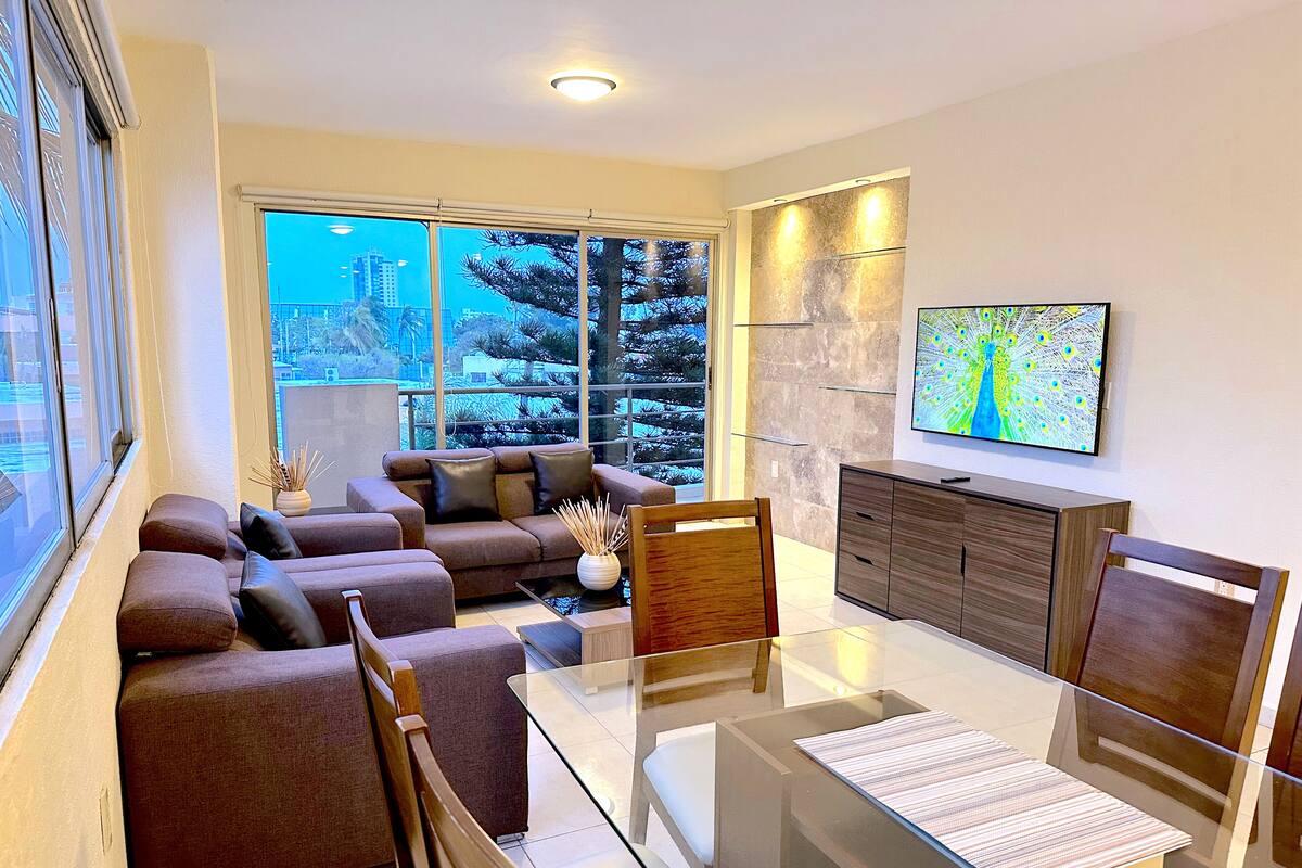 A modern living room with a glass dining table, brown sofas, and a TV on the wall.