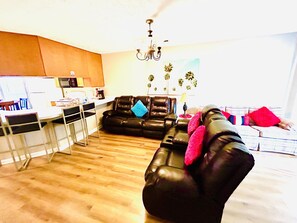 Een moderne woonkamer met een zwarte leren bank, een eethoek met een witte tafel en stoelen, en een keuken op de achtergrond.