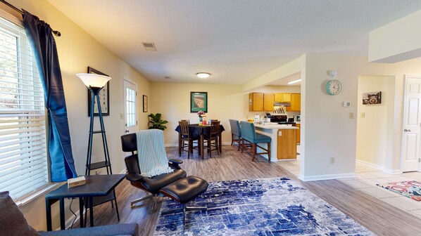 A living room with a blue patterned rug, a dining area with a table and chairs, and a kitchen with wooden cabinets and a stove.
