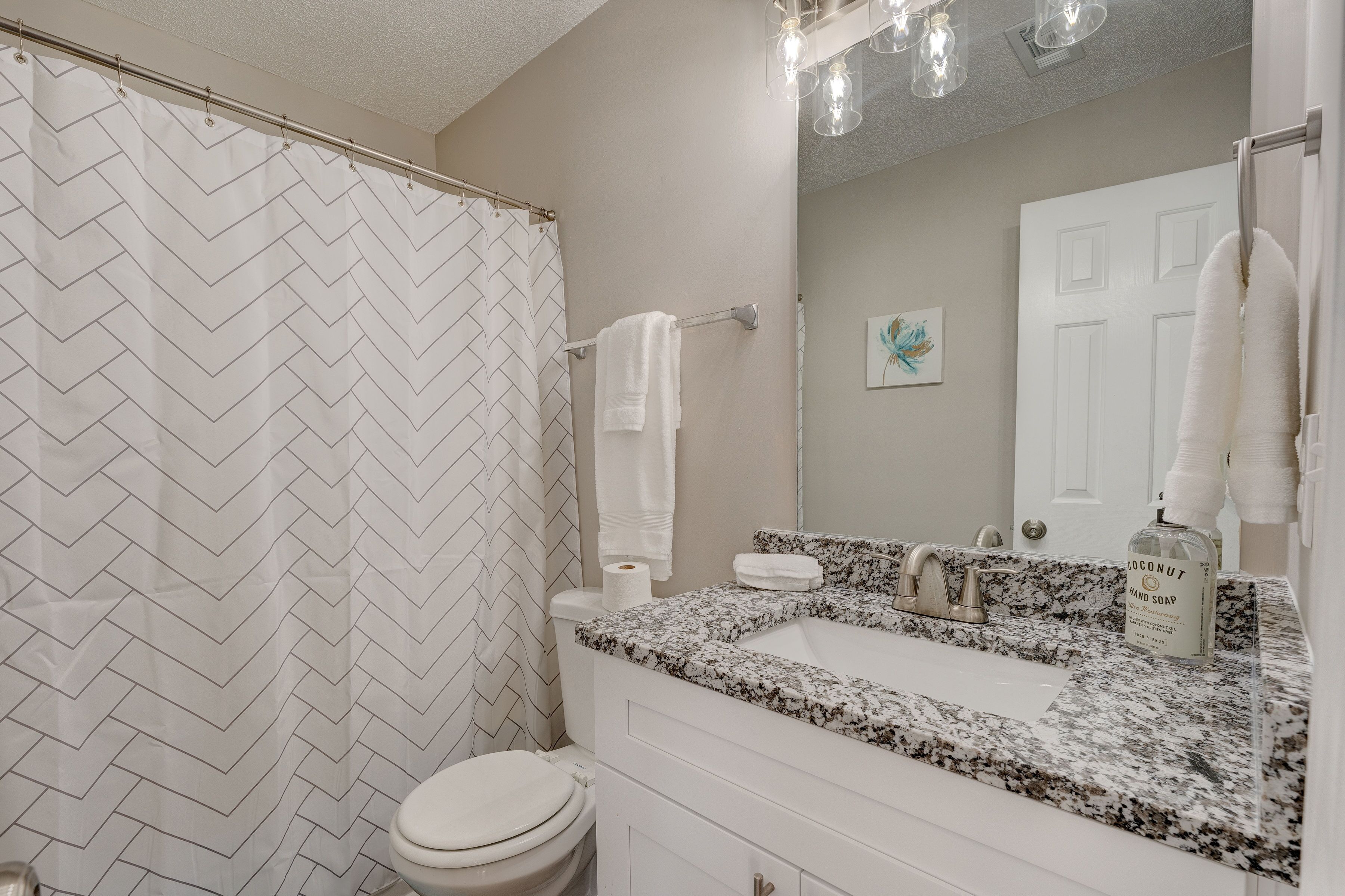A bathroom with a herringbone patterned shower curtain, a toilet, a sink with a granite countertop, and a mirror with a towel rack.