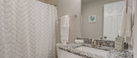 A bathroom with a herringbone patterned shower curtain, a toilet, a sink with a granite countertop, and a mirror with a towel rack.