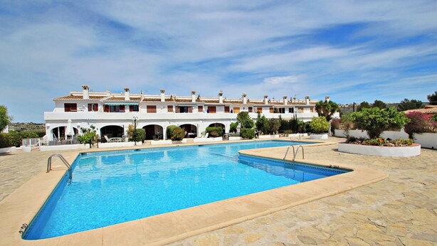 A swimming pool with a stone deck, a white building with multiple balconies, and a clear blue sky. - Jane - modern, well-equipped bungalow in Moraira (Teulada)