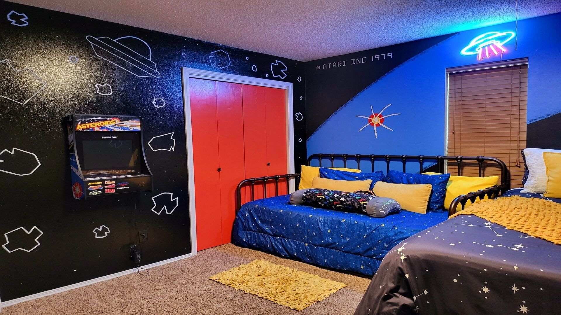 Une chambre avec un mur décoré sur le thème de l’espace, une porte rouge, un lit avec une literie bleue et jaune, et un panneau néon en forme d’OVNI.