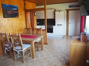 Un intérieur en bois avec une table et des chaises, un coin cuisine avec des meubles blancs et une horloge au mur.